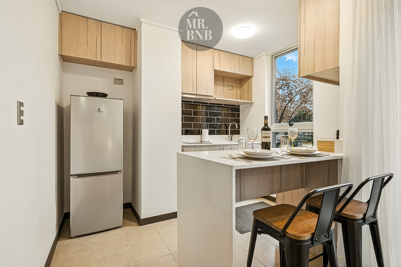 Cocina con refrigerador, mesón, banquetas altas y detalle de vajilla con botella de vino