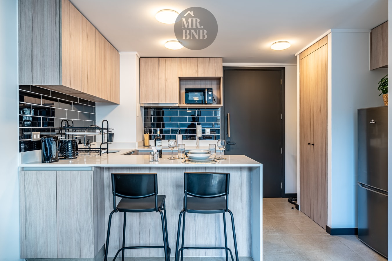 Cocina en U con muebles de madera clara, mesón de piedra, microondas empotrado, refrigerador y dos banquetas negras en la barra