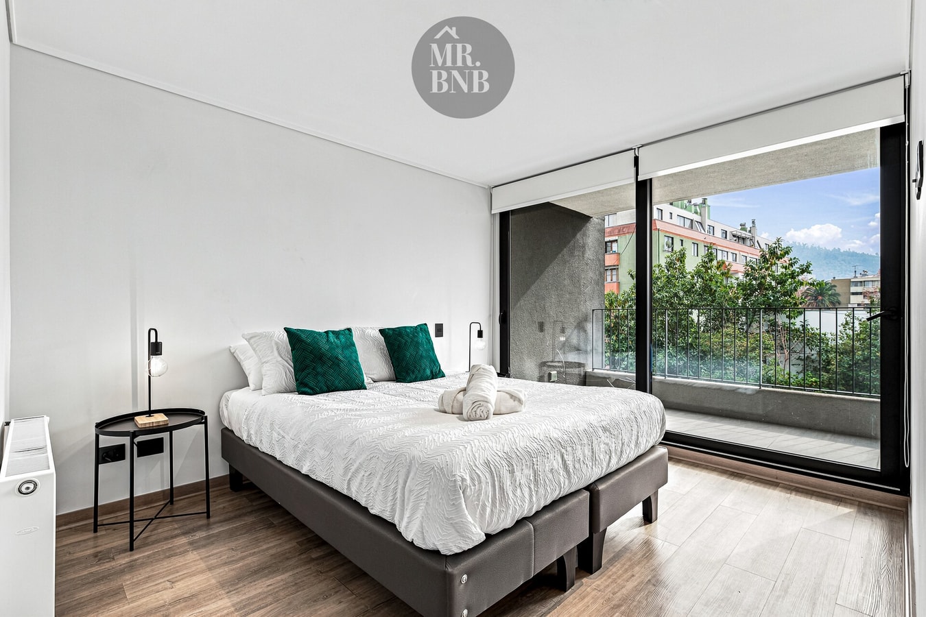 Dormitorio con cama de dos plazas, cojines verdes y ventanal con vista a la ciudad