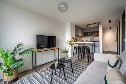 Living con sofá claro, televisor y plantas decorativas, con la cocina visible al fondo