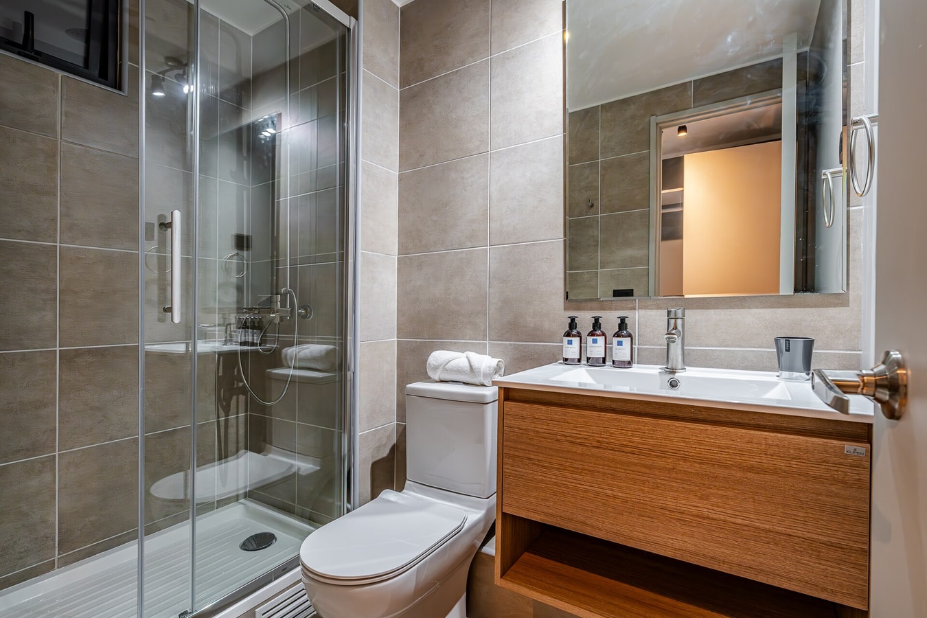 Baño con ducha de vidrio, inodoro y lavamanos de madera con amenities.
