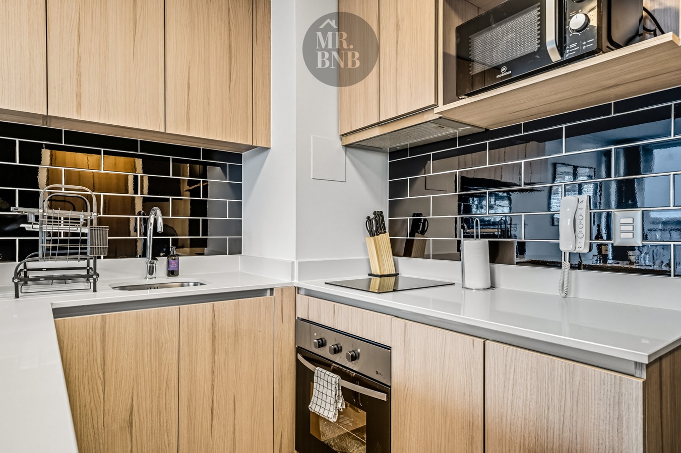 Detalle de la cocina con encimera vitrocerámica, horno, microondas, hervidor, set de cuchillos y citófono en la pared