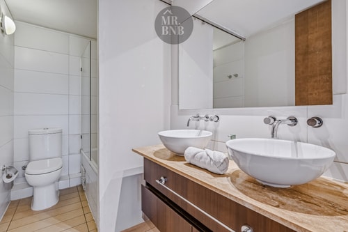 Baño con dos lavamanos tipo bowl sobre mesón de madera, espejo amplio e inodoro.