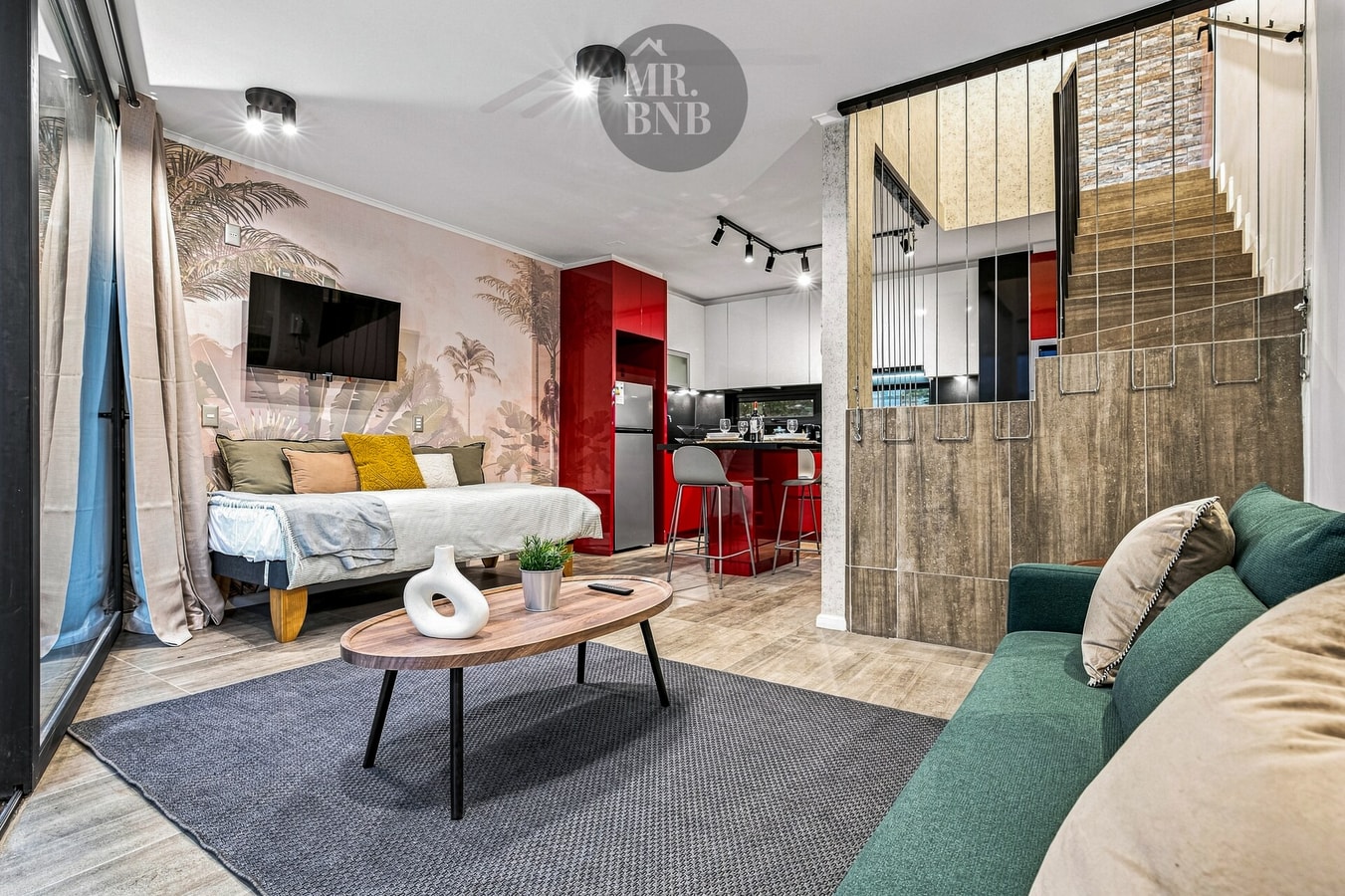 Living con sofá verde y cojines, mesa de centro ovalada, cocina roja al fondo y escalera