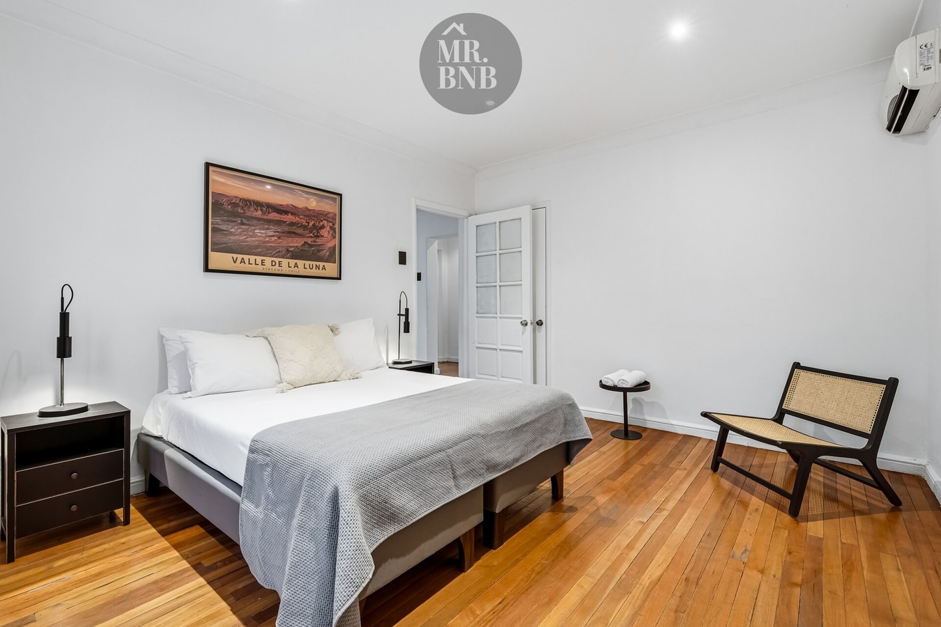 Dormitorio con cama de dos plazas, cuadro, velador y silla de mimbre.