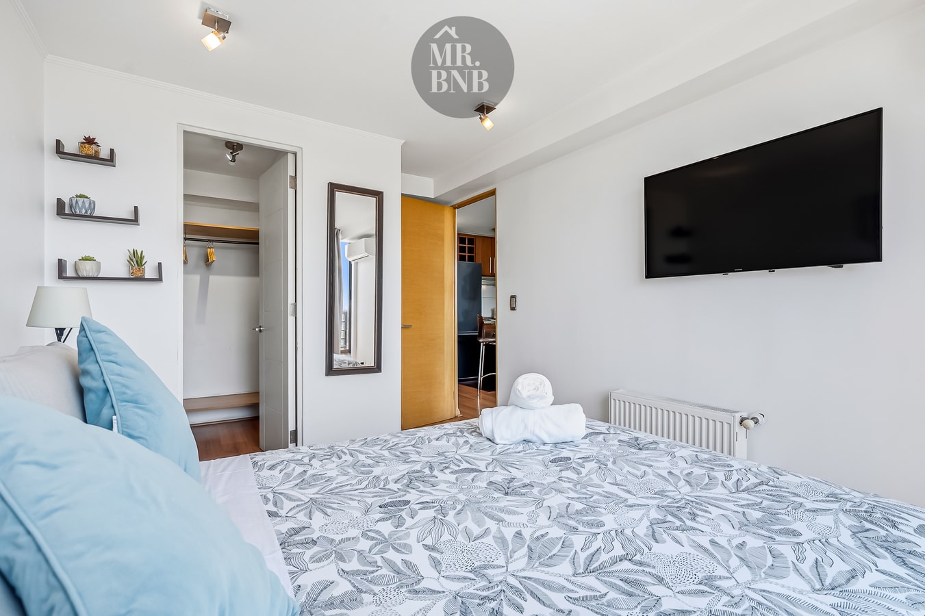 Dormitorio con cama, repisas decorativas, televisor y ventanal con cortinas.