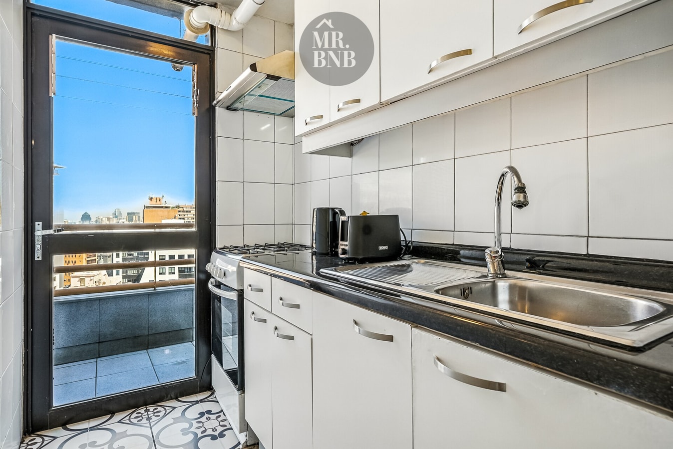 Cocina con lavaplatos, muebles blancos y puerta hacia balcón con vista a la ciudad
