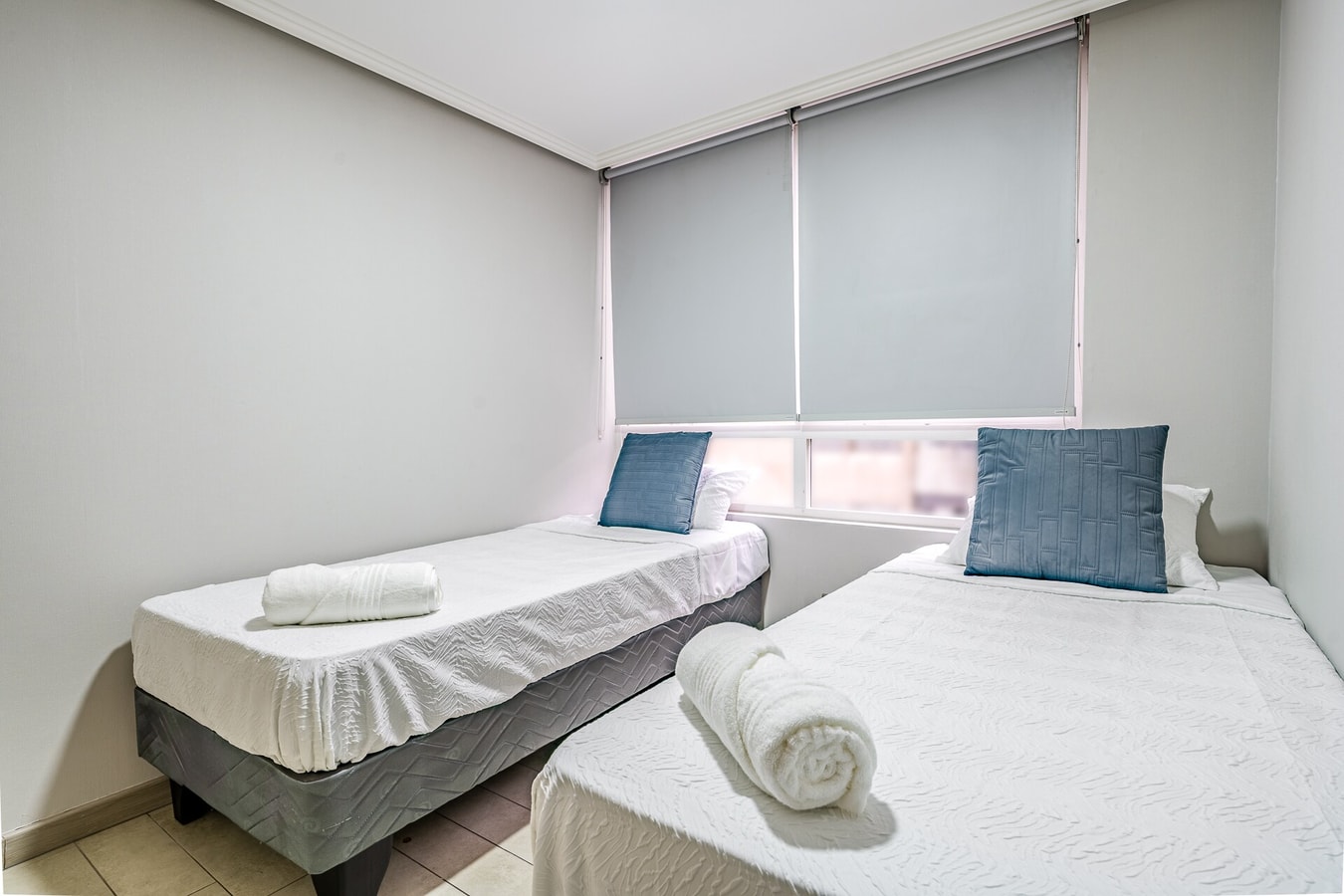 Dormitorio con dos camas individuales, cojines azules y persiana enrollable.