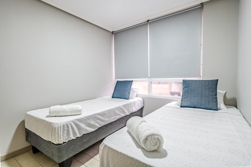 Dormitorio con dos camas individuales, cojines azules y persiana enrollable.