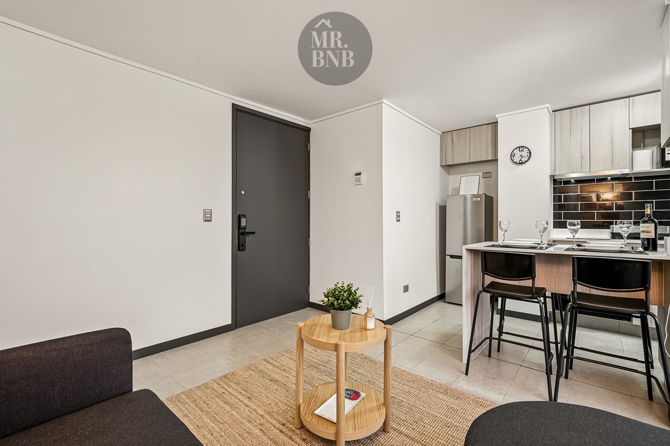 Hall de entrada con puerta de acceso oscura y cocina con barra al fondo.