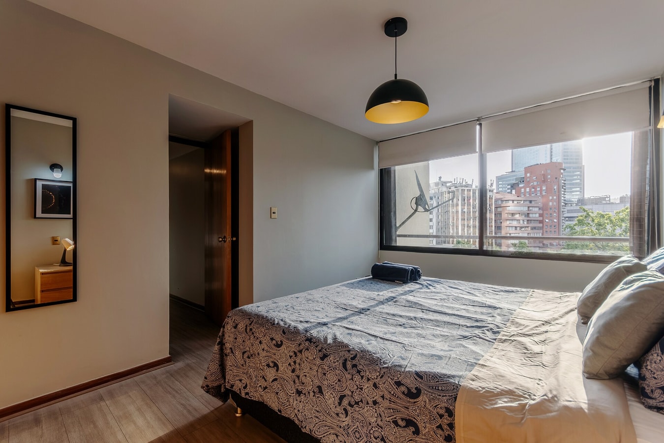 Dormitorio con cama matrimonial, espejo y ventana con vista a edificios.