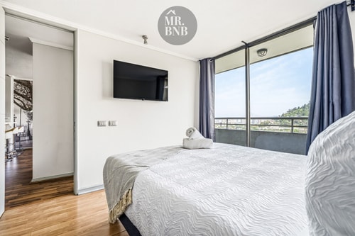 Dormitorio con cama, televisor en la pared y ventanal con cortinas.
