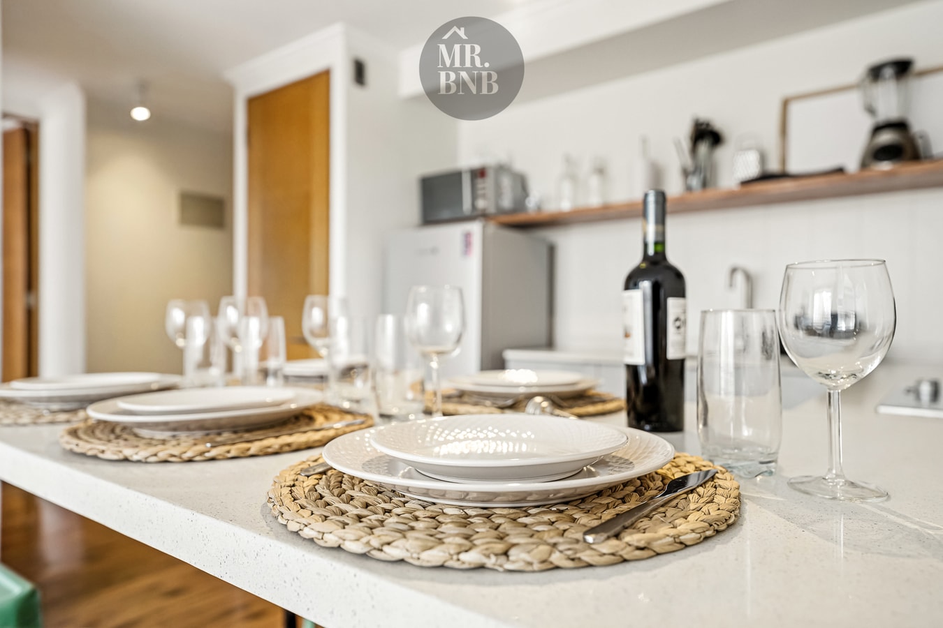 Comedor con mesa puesta, platos, copas y botella de vino, cocina al fondo