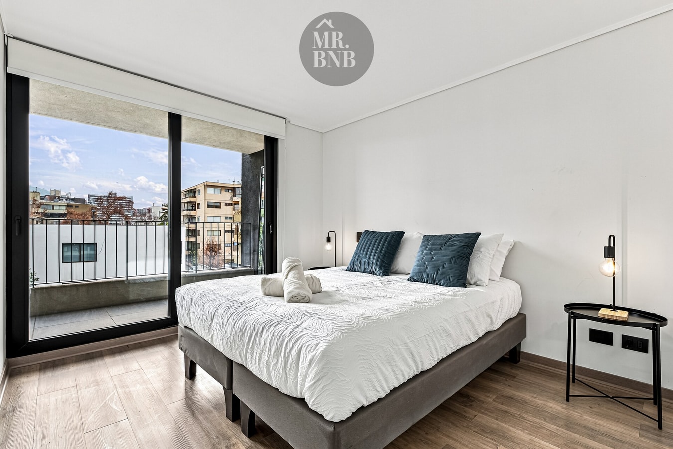 Dormitorio con cama de dos plazas, cojines verdes, ventanal con vista urbana y velador
