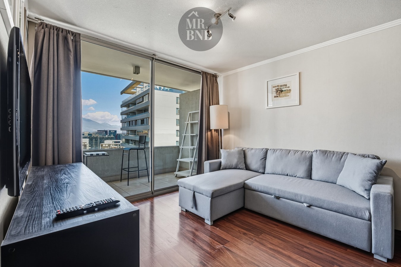 Living con sofá esquinero gris, televisor y ventanal hacia edificios