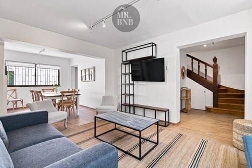 Living con sofá, mesa de centro y Smart TV en mueble, junto a la escalera de madera que sube al segundo piso.
