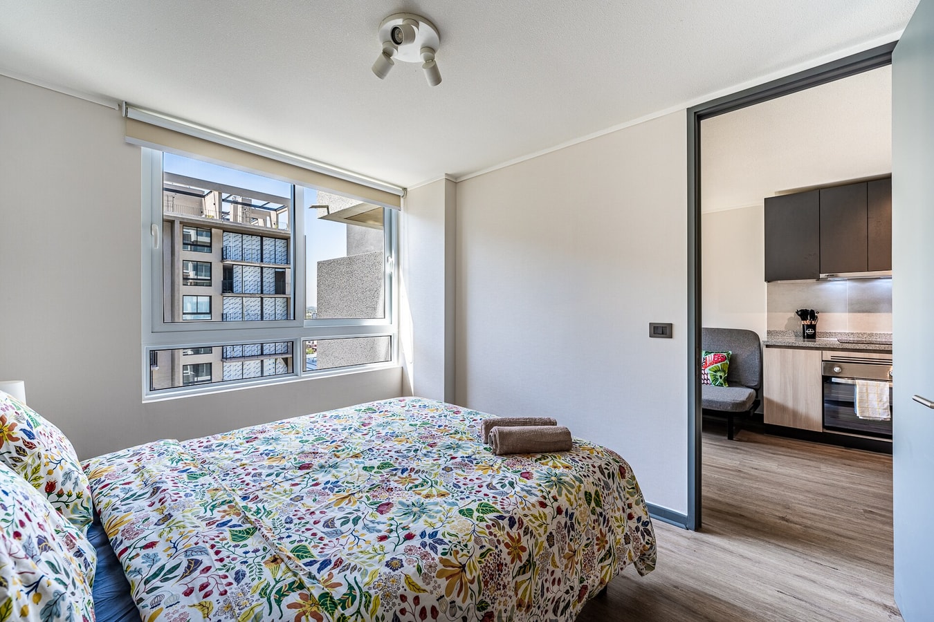 Dormitorio con dos camas individuales con cobertor floral, ventana con vista a edificios y puerta corrediza hacia la cocina.