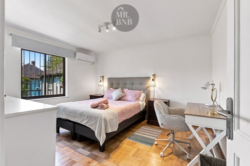 Dormitorio con cama matrimonial, veladores con lámparas, escritorio con silla, aire acondicionado y ventana a los árboles.