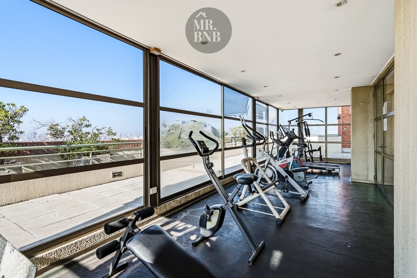 Gimnasio del edificio con bicicletas, elíptica y banco de pesas junto a ventanales panorámicos