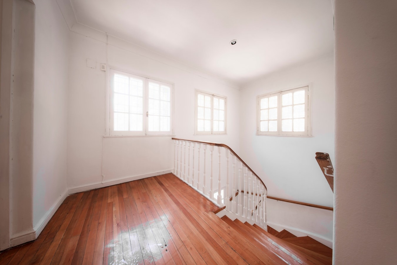 Habitación vacía con piso de madera, tres ventanas de vidrio repartido y la escalera de balaustres blancos al costado