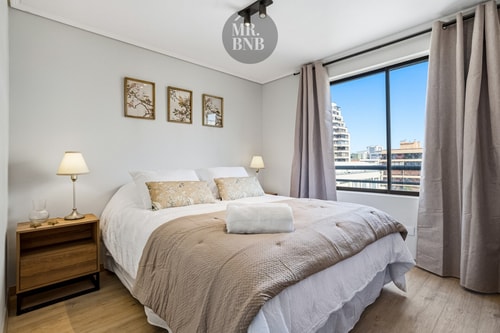 Dormitorio con cama matrimonial junto a ventana con vista a edificios, cuadros sobre el respaldo
