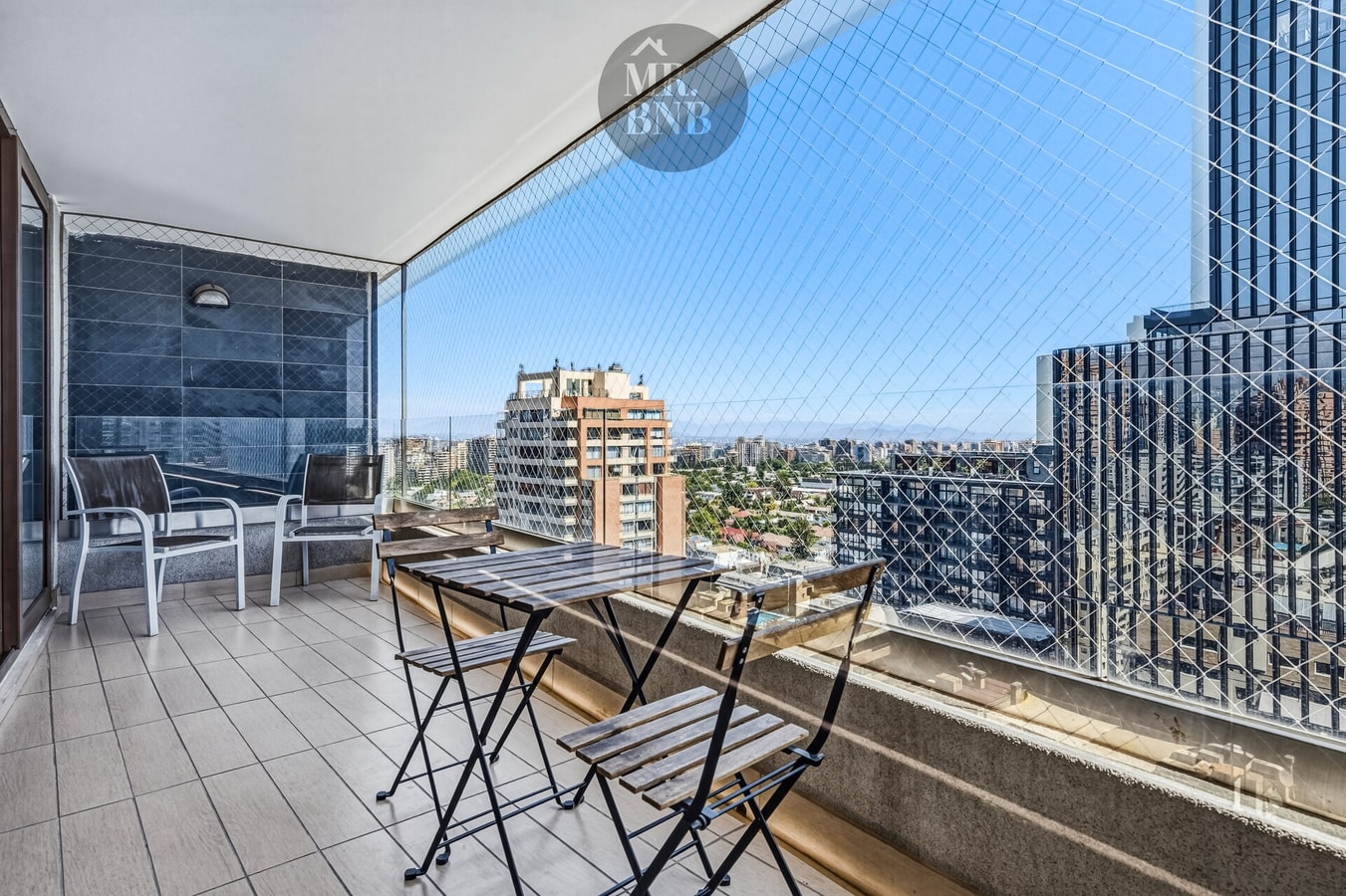 Terraza con mesa y dos sillas, malla de seguridad y vista panorámica de la ciudad.
