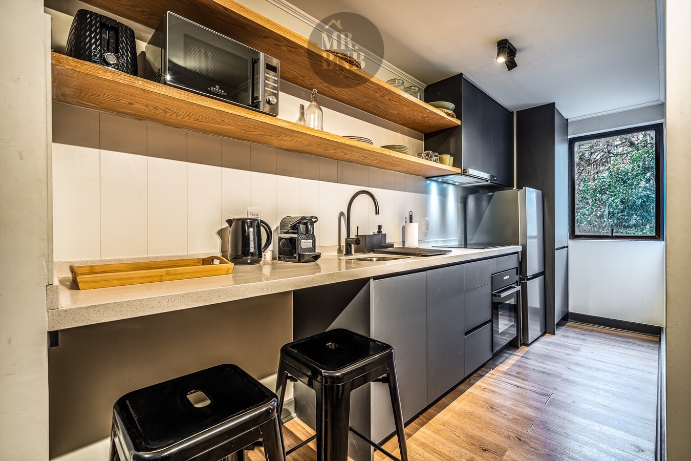 Cocina con encimera larga, muebles negros, microondas, hervidor, cafetera y refrigerador