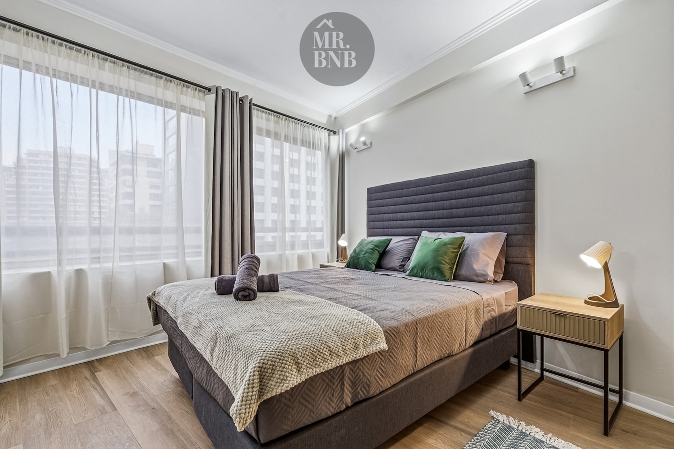 Dormitorio con cama, veladores, cortina y ventanal con vista a edificios