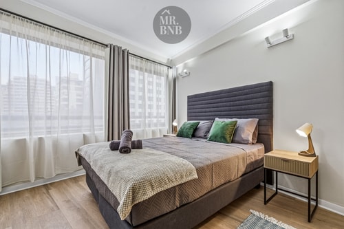 Dormitorio con cama, veladores, cortina y ventanal con vista a edificios