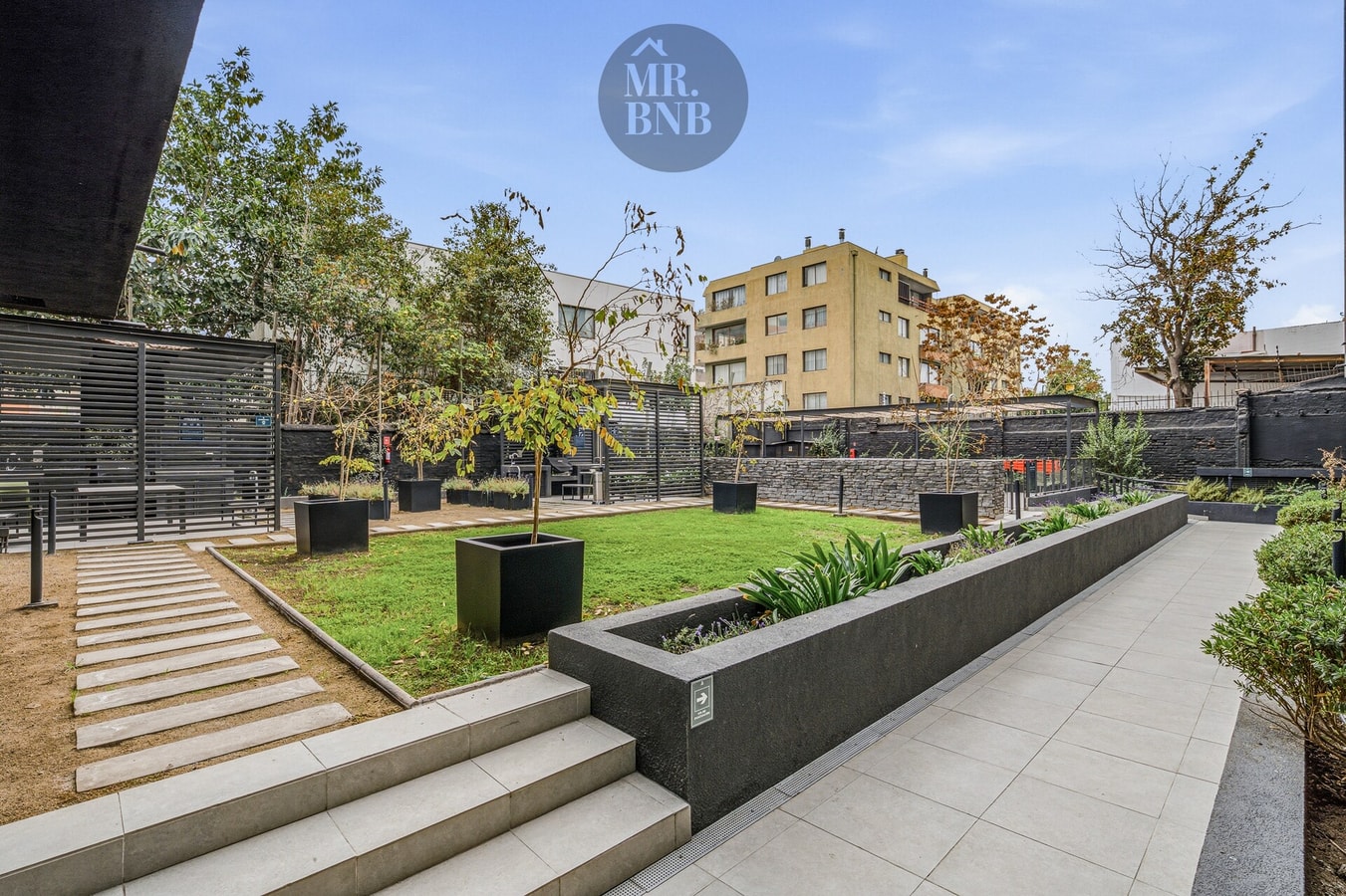 Jardín interior del edificio con senderos de piedra, pasto y arbustos junto a edificios del entorno