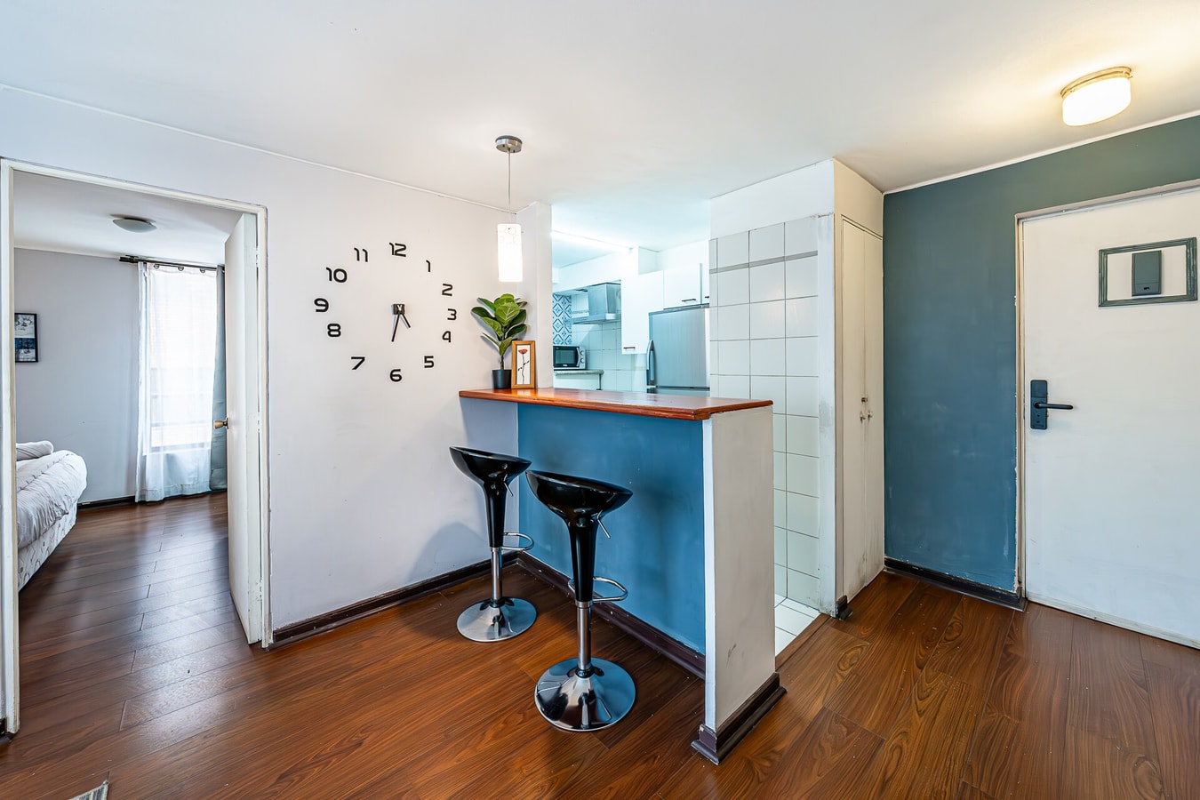Living comedor con barra de madera y dos banquetas negras, reloj de pared grande, muro azul, puerta de acceso y el dormitorio al fondo