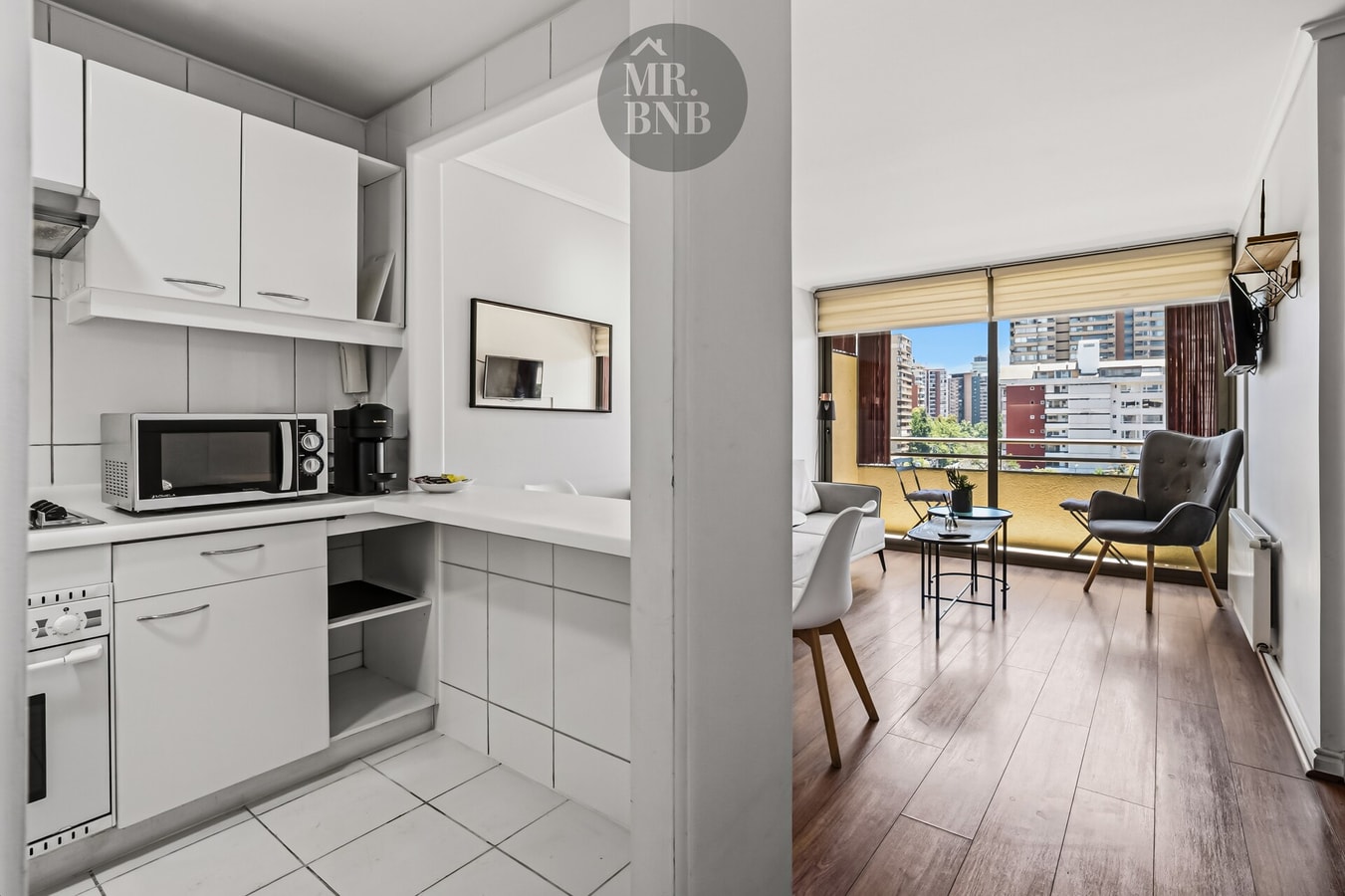 Cocina blanca en L con microondas, living con mesa y sillas al fondo.