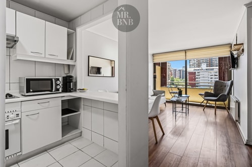 Cocina blanca en L con microondas, living con mesa y sillas al fondo.