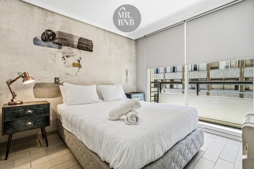 Dormitorio con cama, veladores rústicos y ventanal con vista a edificios