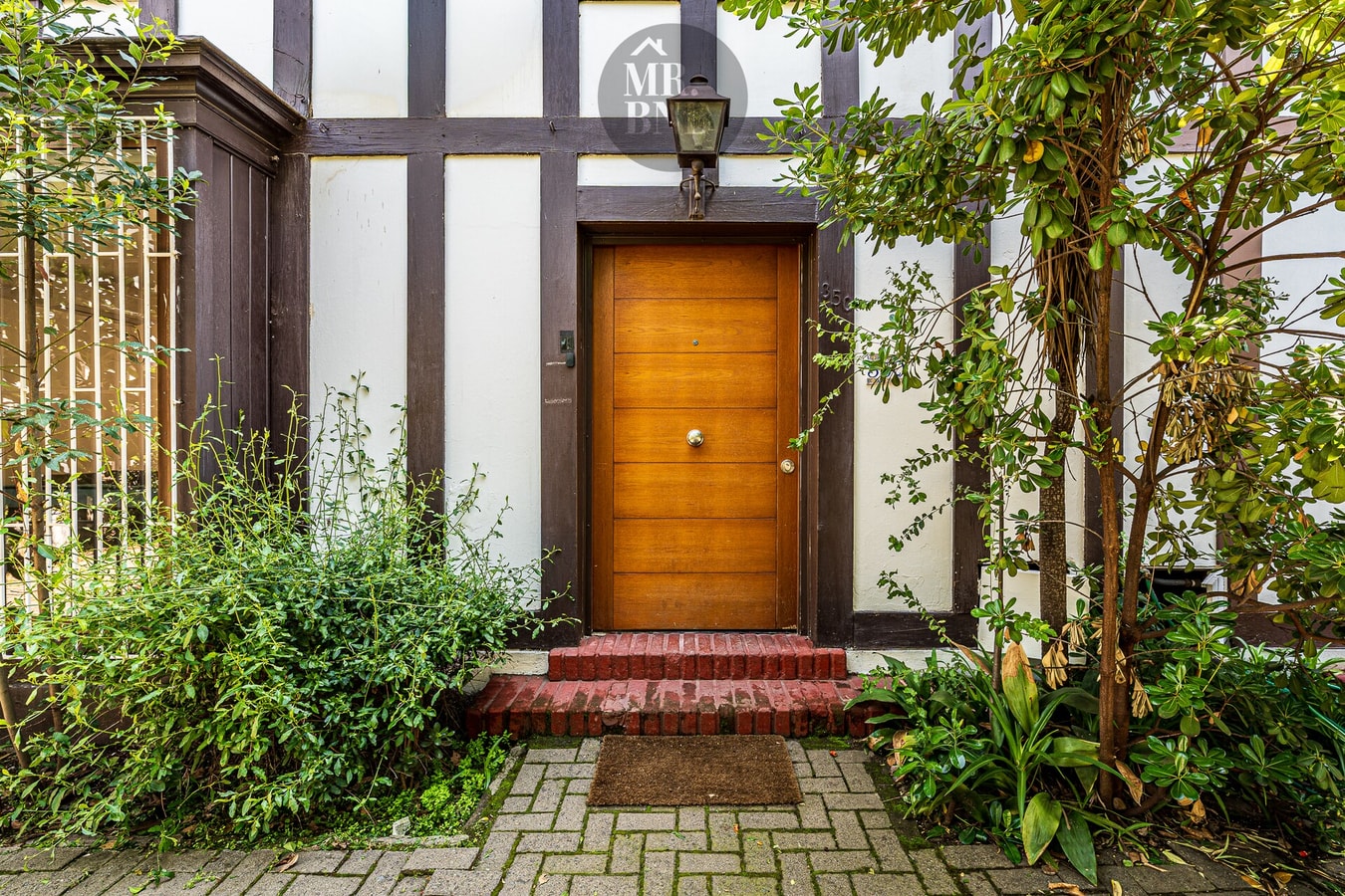 Puerta de entrada de madera con farol, escalones de ladrillo y jardín.