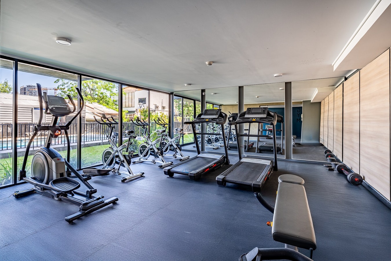 Gimnasio del edificio con máquinas de cardio, banco de pesas y ventanales hacia el jardín.