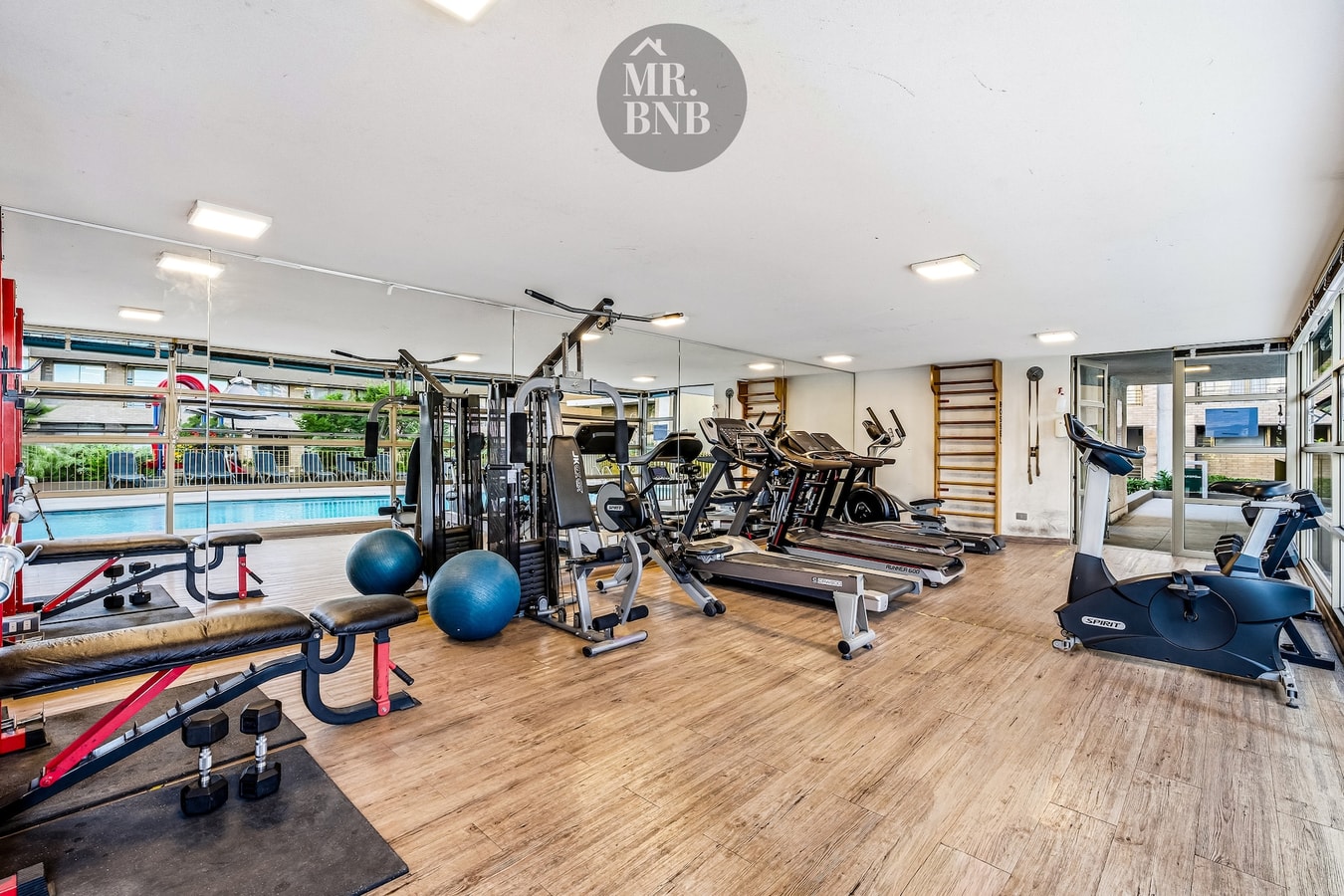 Gimnasio del edificio con máquinas de musculación, trotadoras y balones, con vista a la piscina