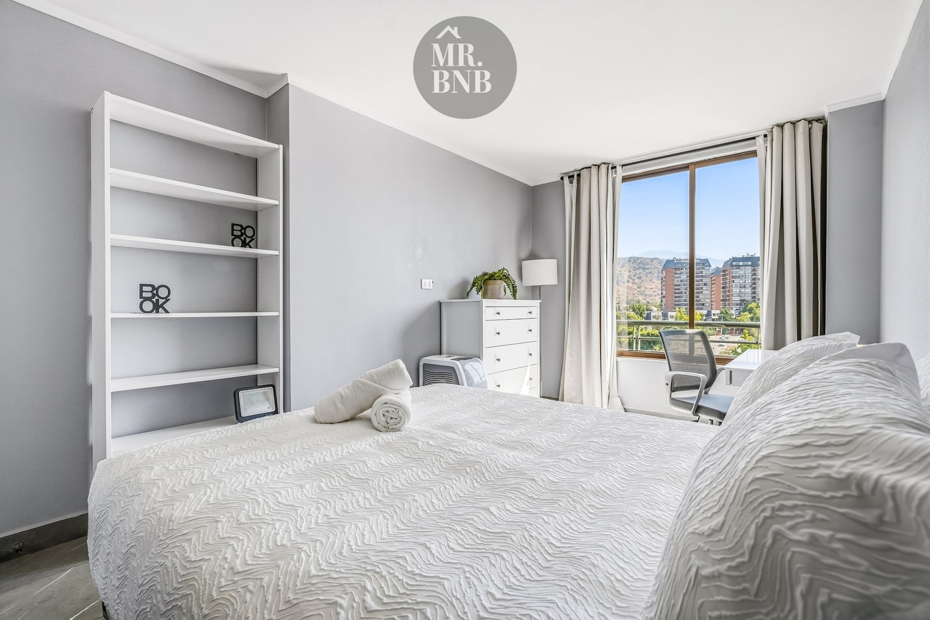 Dormitorio con cama, repisas blancas, cómoda y ventana con vista a la ciudad