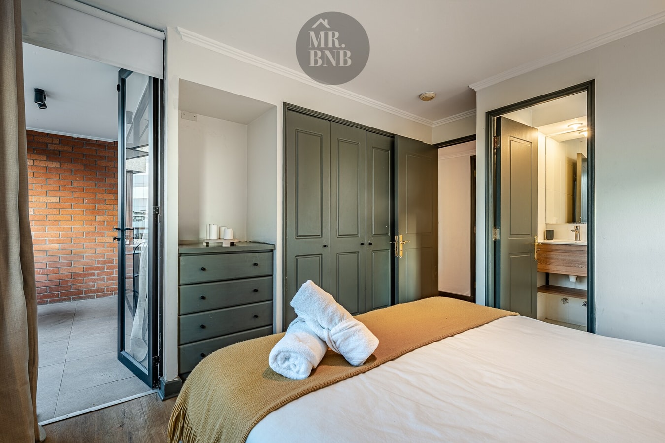 Dormitorio con cómoda de madera oscura, cama con pie de cama mostaza y toallas dobladas