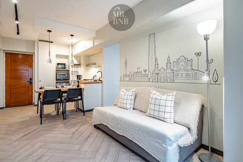 Living con sofá cama blanco y cojines, mural con dibujo de la ciudad y comedor al fondo