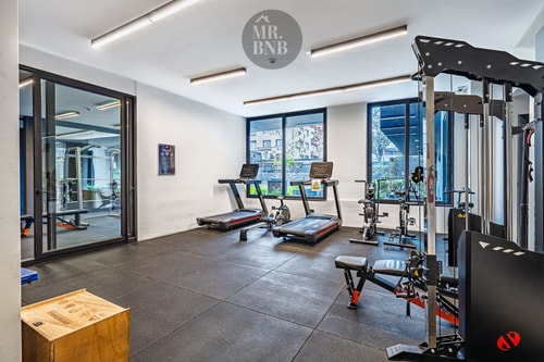 Gimnasio del edificio equipado con máquinas de cardio y musculación, piso de goma y ventanales.