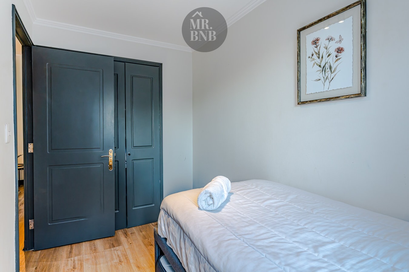 Rincón de dormitorio con puertas de clóset oscuras, cuadro floral y borde de cama con toallas