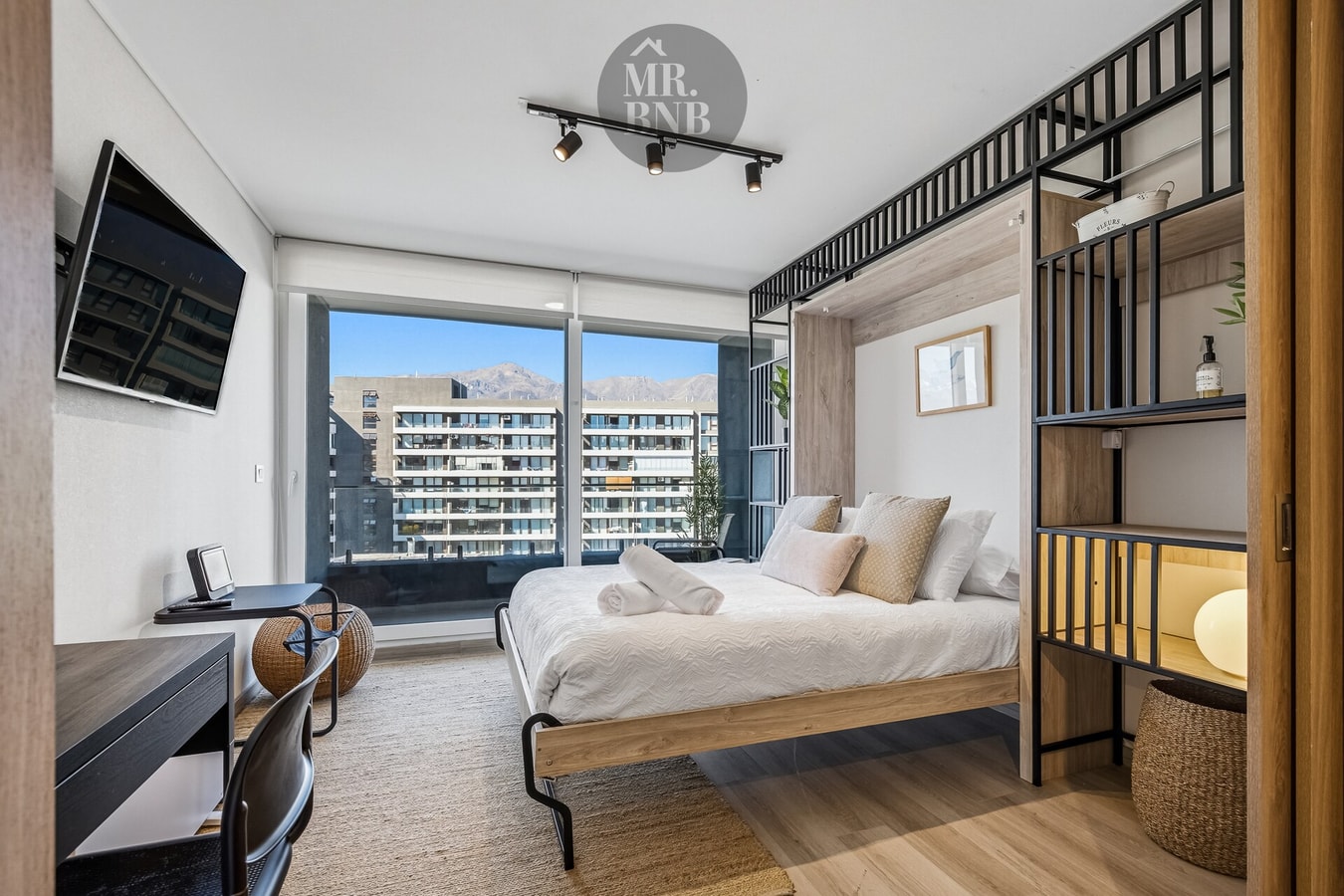 Estudio con cama Murphy desplegada, TV, escritorio y ventanal con vista a la ciudad