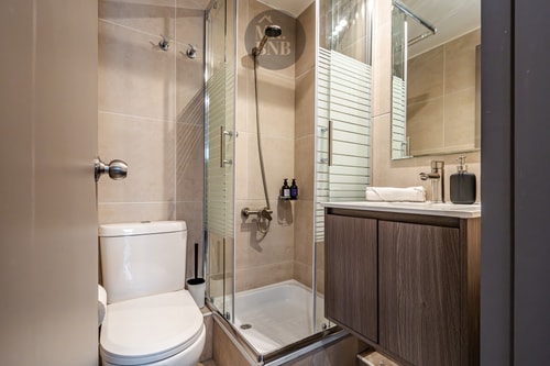 Baño con ducha de vidrio, vanitorio de madera e inodoro.