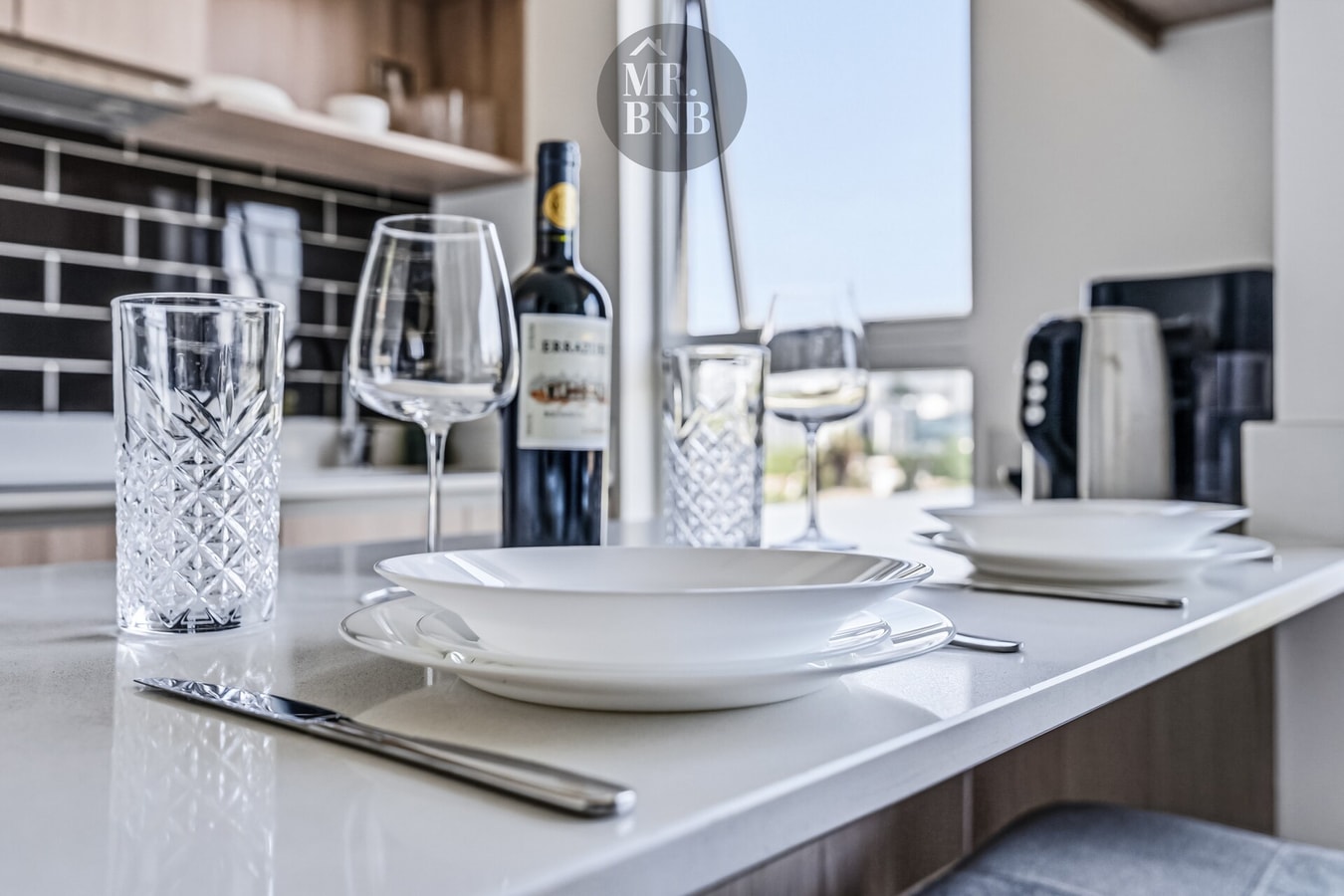 Detalle de mesa puesta con vajilla, copas y botella de vino sobre encimera de cocina