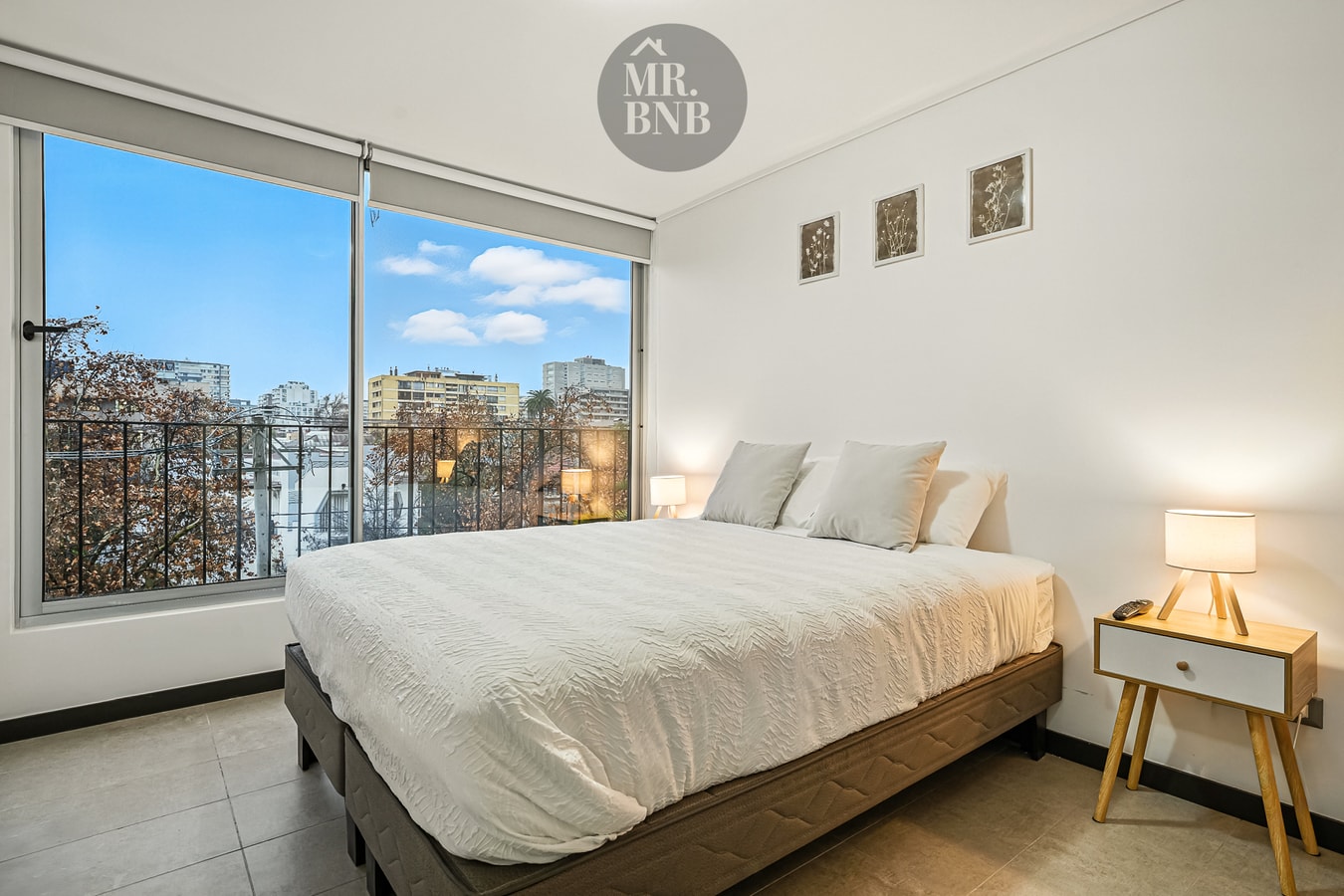Dormitorio con cama, velador con lámpara y ventanal con vista a la ciudad.