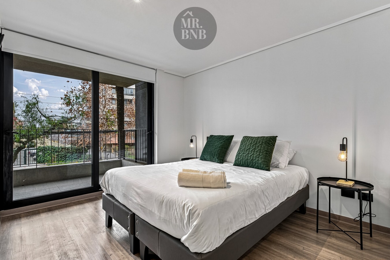 Dormitorio con cama de dos plazas, cojines verdes y ventanal con vista a la calle.