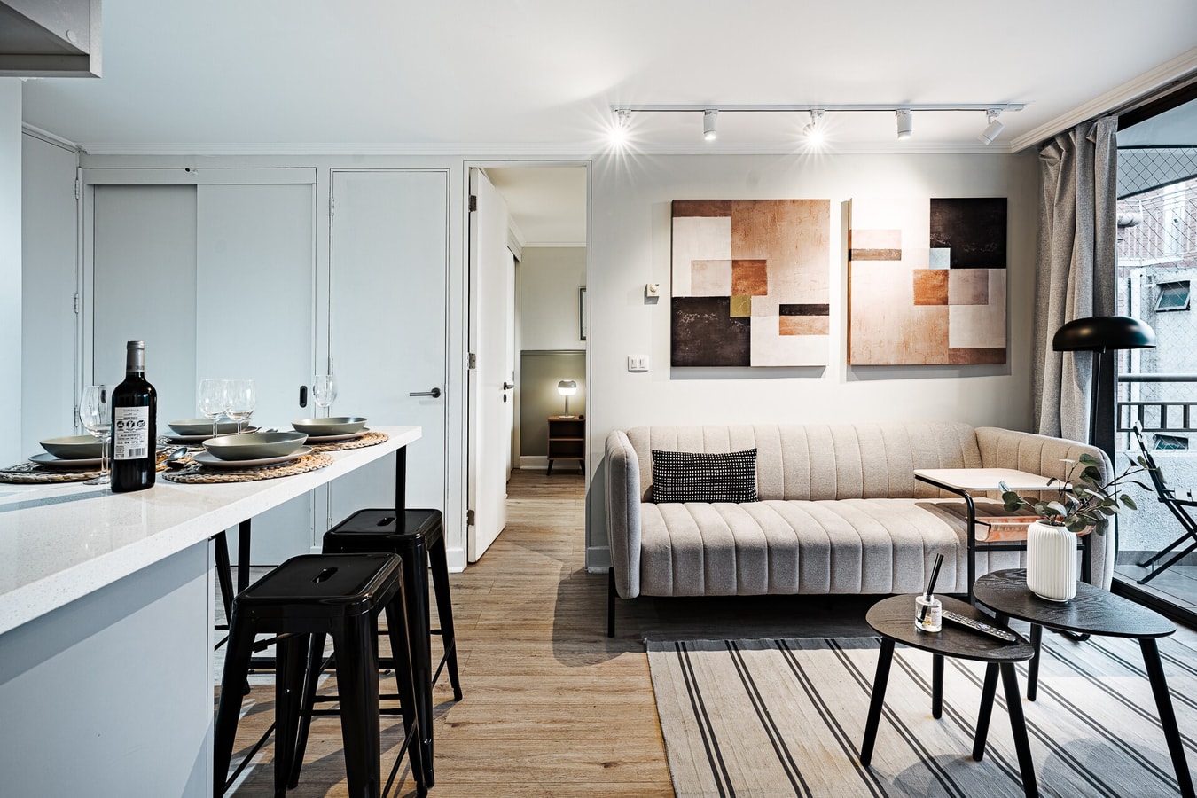 Cocina comedor con barra, banquetas negras y living con sillón y cuadros al fondo
