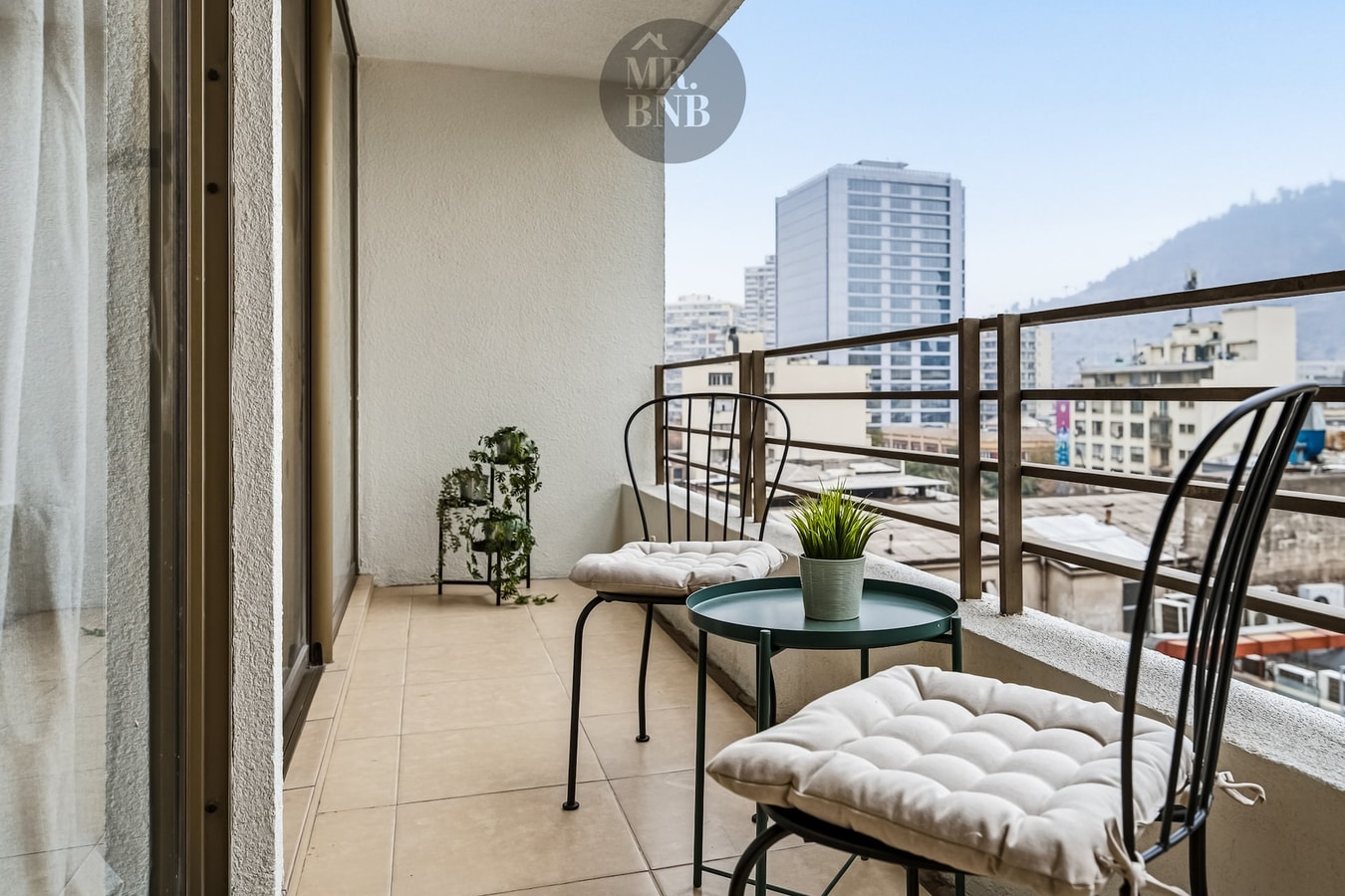 Terraza privada con mesa, sillas y plantas, con vista a edificios y cerro