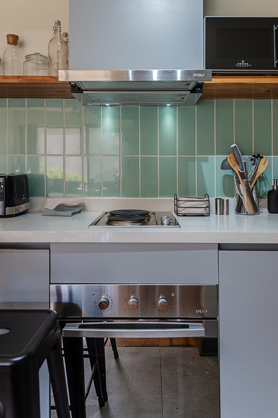 Cocina con encimera y horno de acero inoxidable bajo la campana, azulejo verde agua y utensilios sobre el mesón